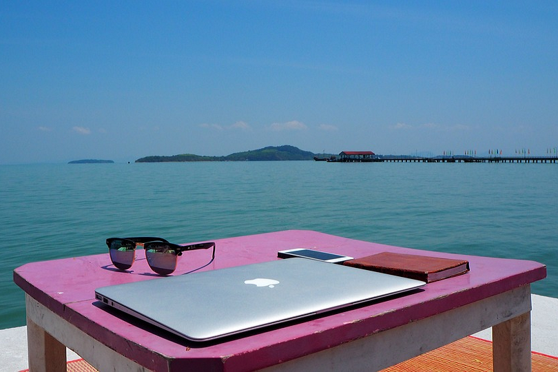 Óculos, iphone e macbook em uma mesa de frente para o mar - Blog Viva de Pousada